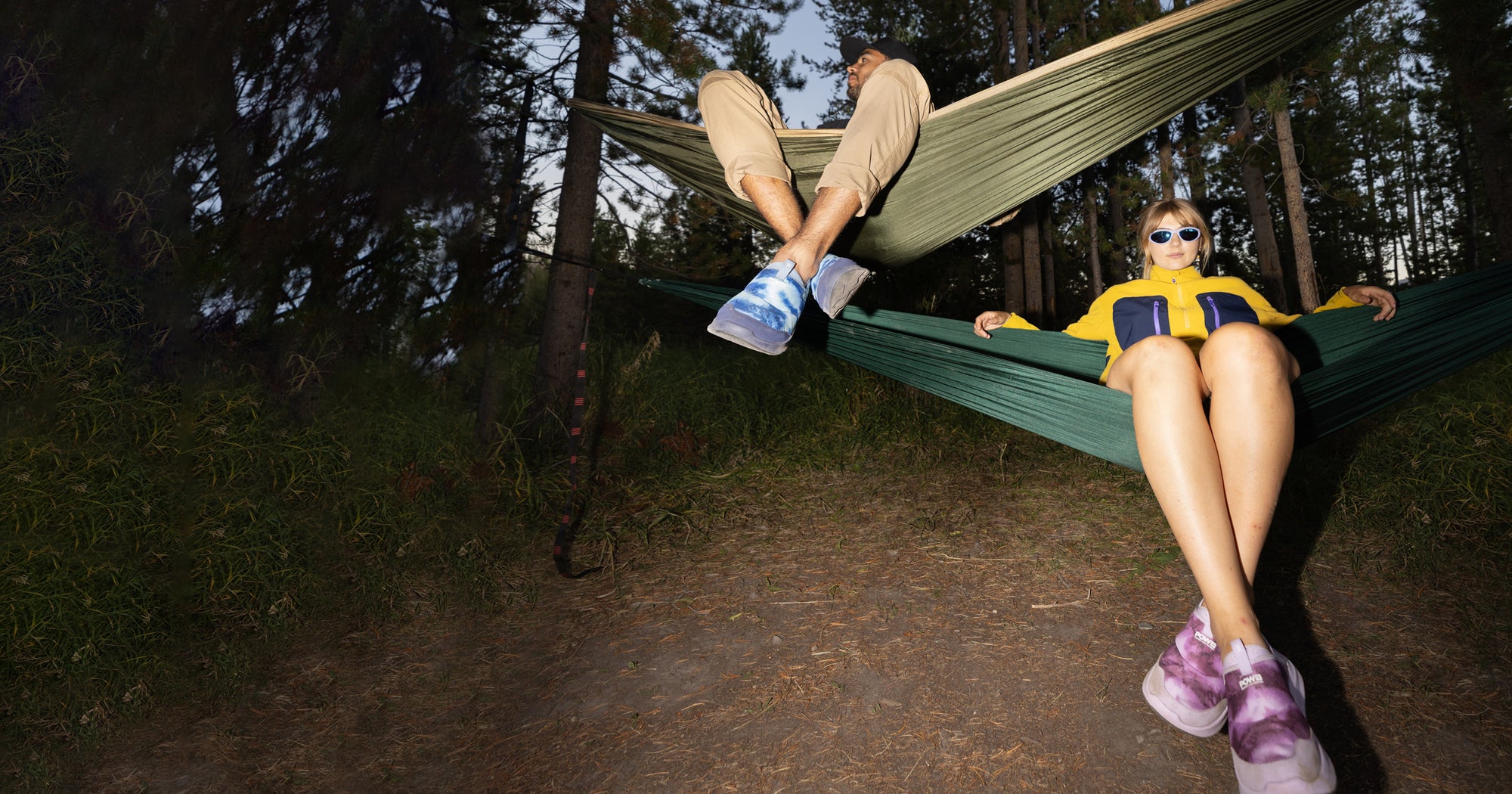 A man and a woman sit comfortably in a hammock, surrounded by nature, sharing a serene moment while both wearing bright colored slip-on shoes.