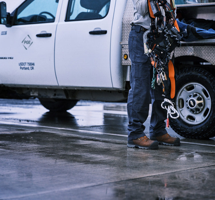 A person standing next to a truck, holding a large tool belt/harness and wearing a pair of brown KEEN Utility work boots.
