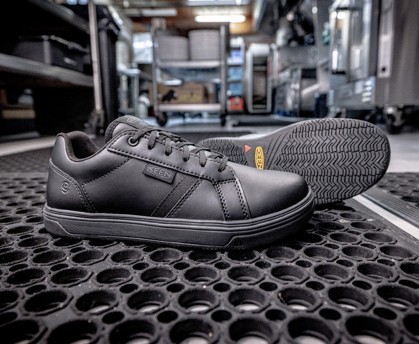 Product shot of black, leather work sneakers on floor of restaurant kitchen, showcasing waterproofness, slip-resistance and cushioning of shoes.