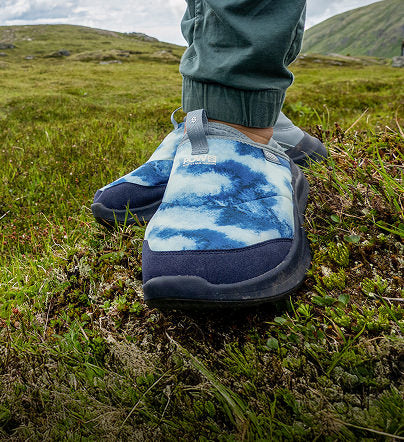 Person positioned on a grassy field, displaying a pair of blue slip-on shoes, surrounded by lush greenery and open space.