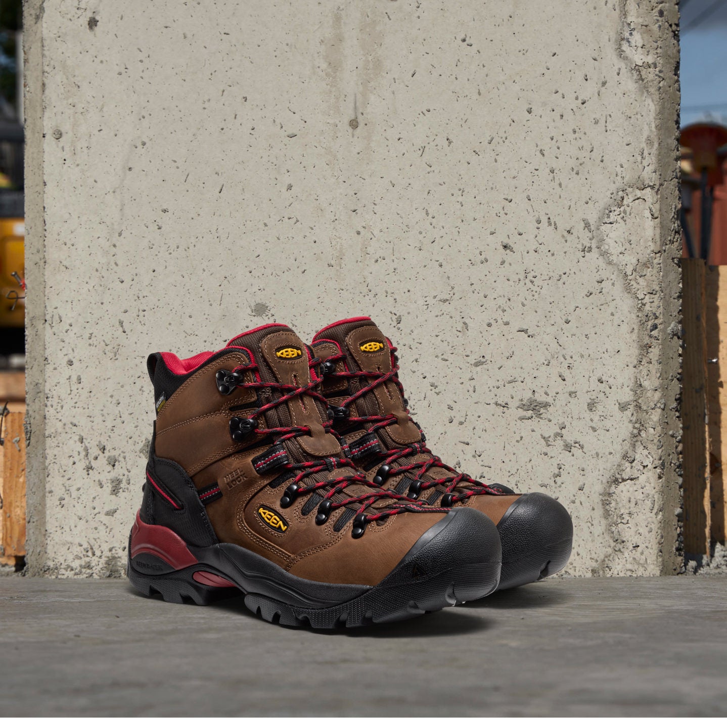 Pair of brown leather work boots with red and black laces and yellow keen logos leaning against a concrete wall.