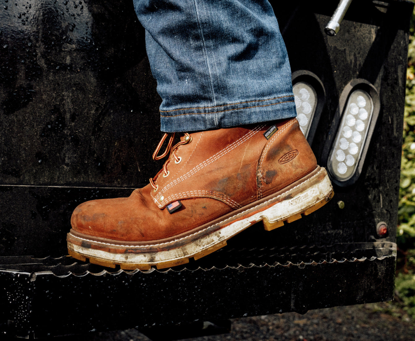 Person wearing worn brown leather work boots featuring a tiny american-flag tag, with jeans stepping onto a truck platform.