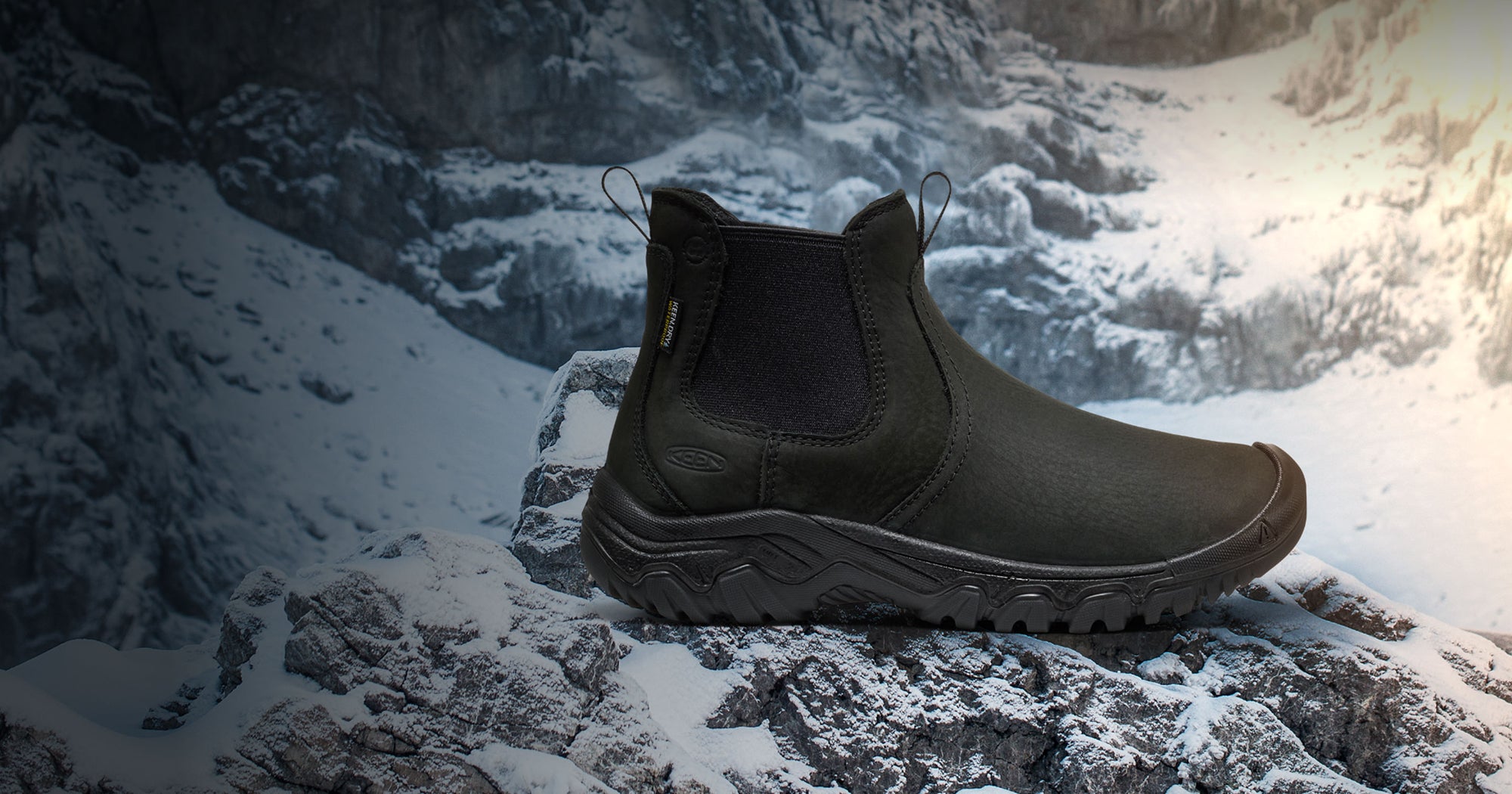 Black leather, insulated slip-on boot with rugged tread and elastic side panels, shown on a snowy rock in the mountains.
