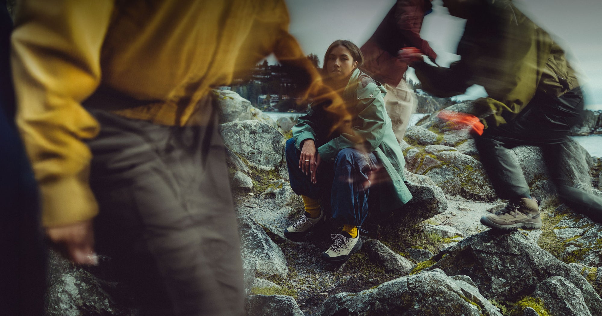 Woman in casual winter clothes sitting on a rocky pier while three other blurry humans move around her.
