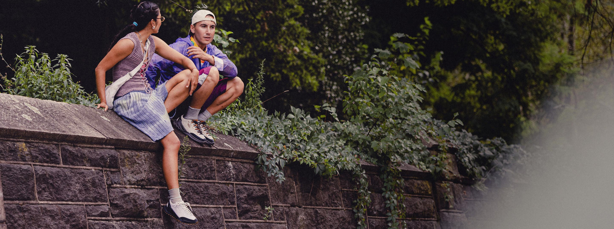 A man and woman sitting casually on a brick wall, enjoying each other's company against a backdrop of nature while wearing Jasper Zionic sneakers.