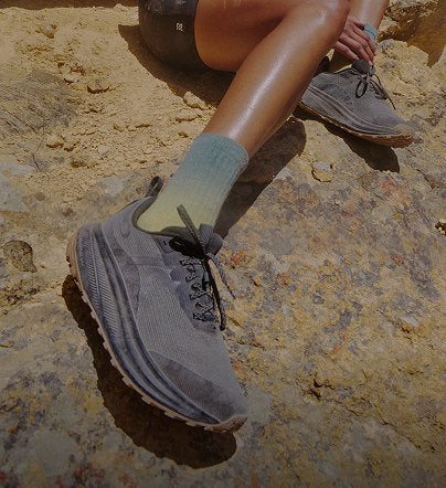 A woman seated on a rock, displaying her grey Roam trail running shoes in an high desert setting.