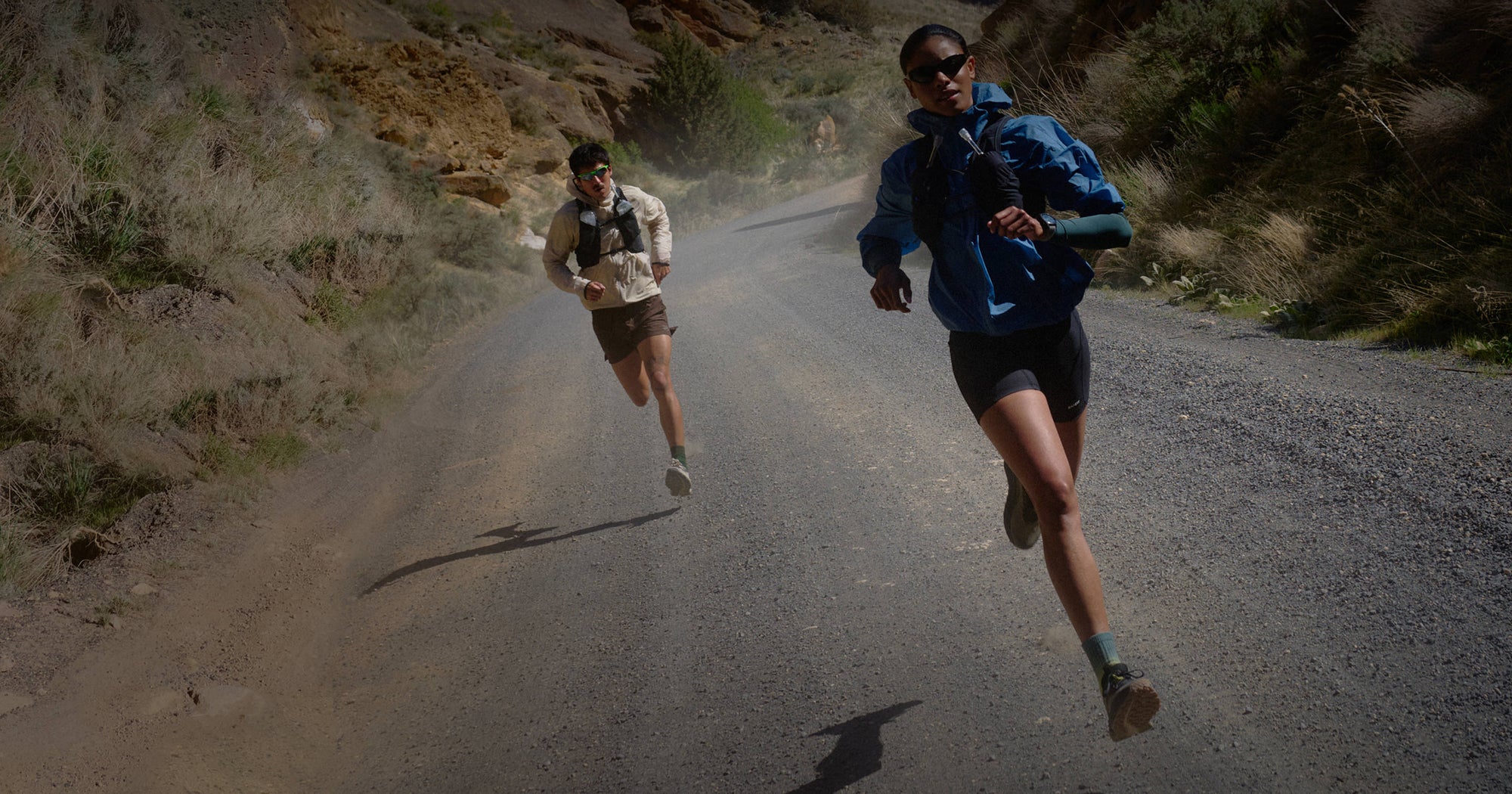 Two people wearing Roam trail running shoes jog on a dirt road, framed by scenic mountain views.