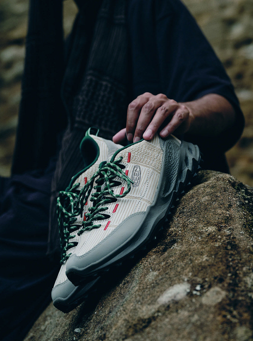 A person on a rock holds sneakers featuring green laces and accent stitching, dressed in casual clothing in outdoor setting.