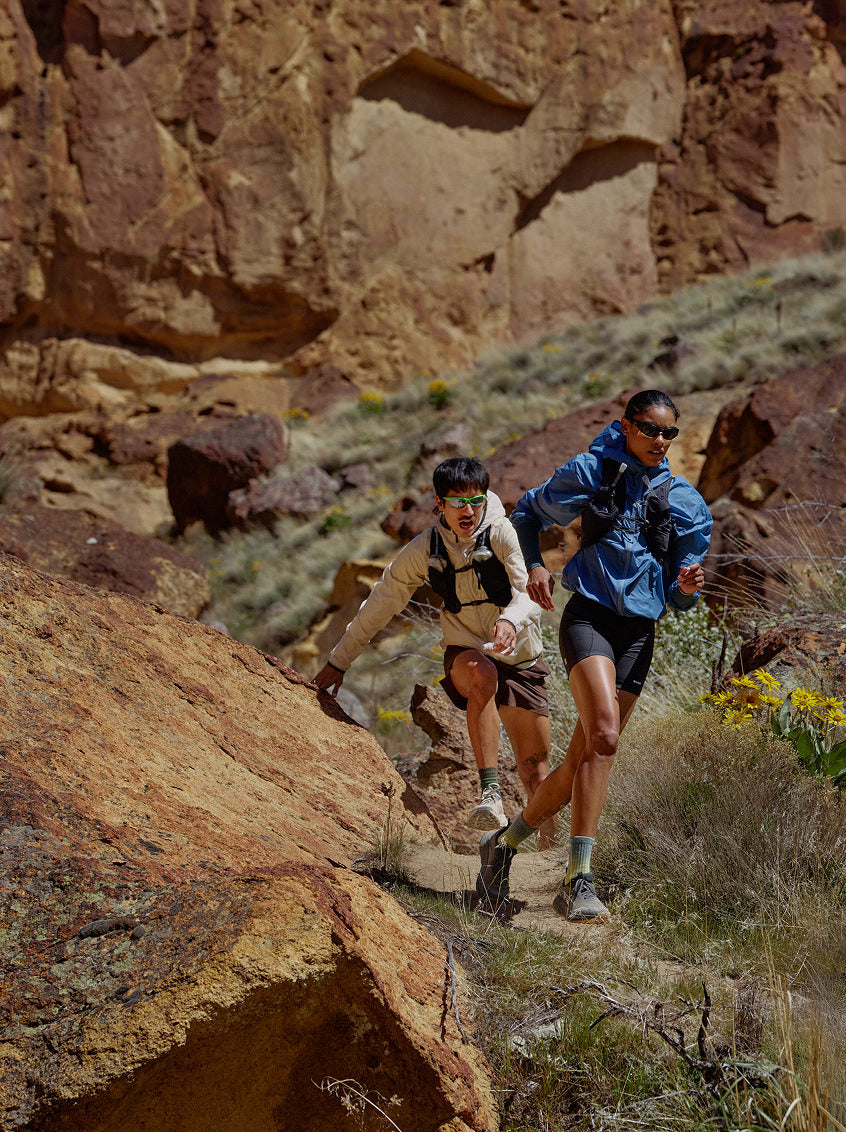 Two individuals running side by side on a dirt trail surrounded by desert plants and sandstone rocks. 