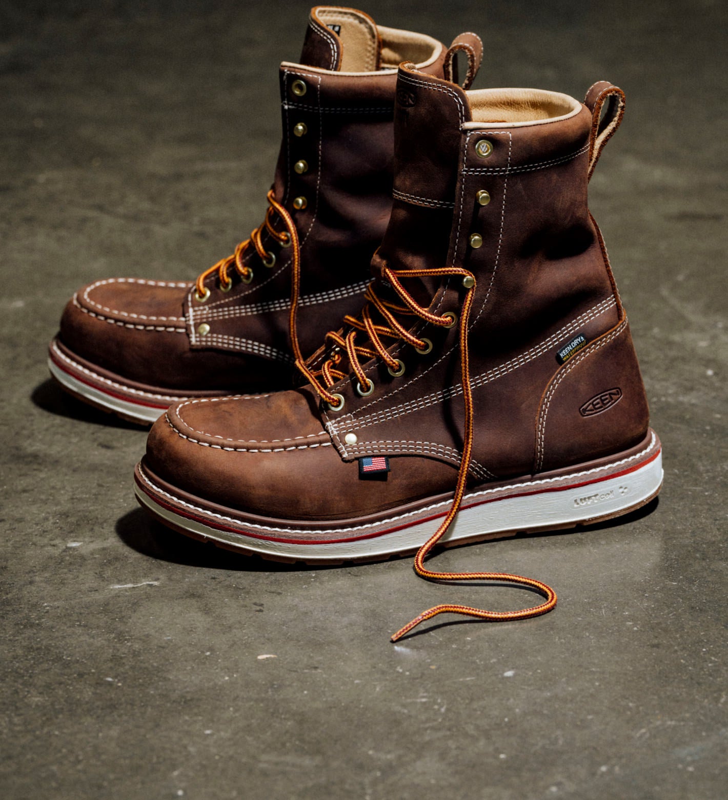 Pair of brown leather work boots with orange laces and U.S. flag tag on a concrete floor.