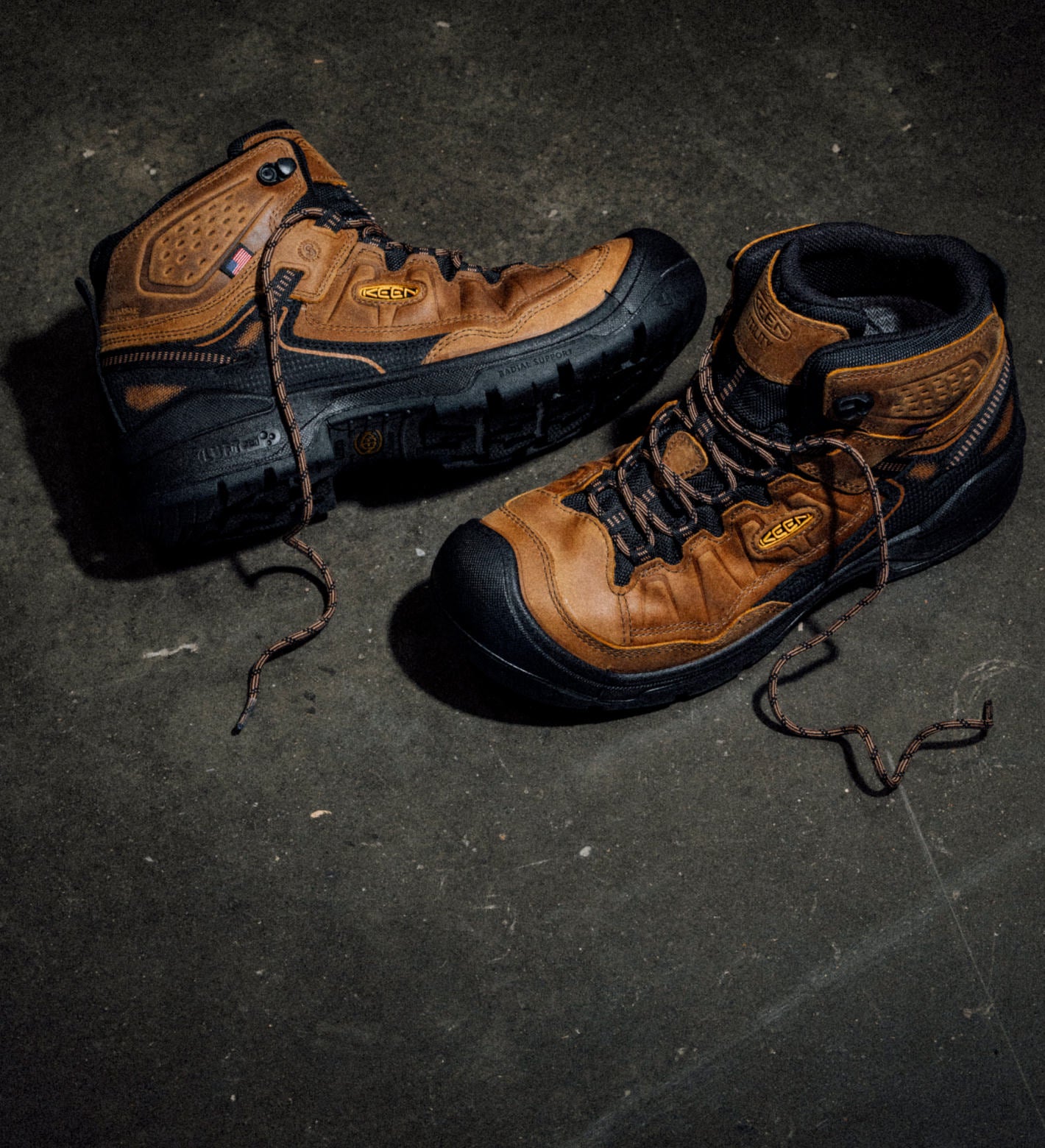 Pair of brown and black mid-cut work boots with a classic hiking-boot design laying on a concrete floor. 