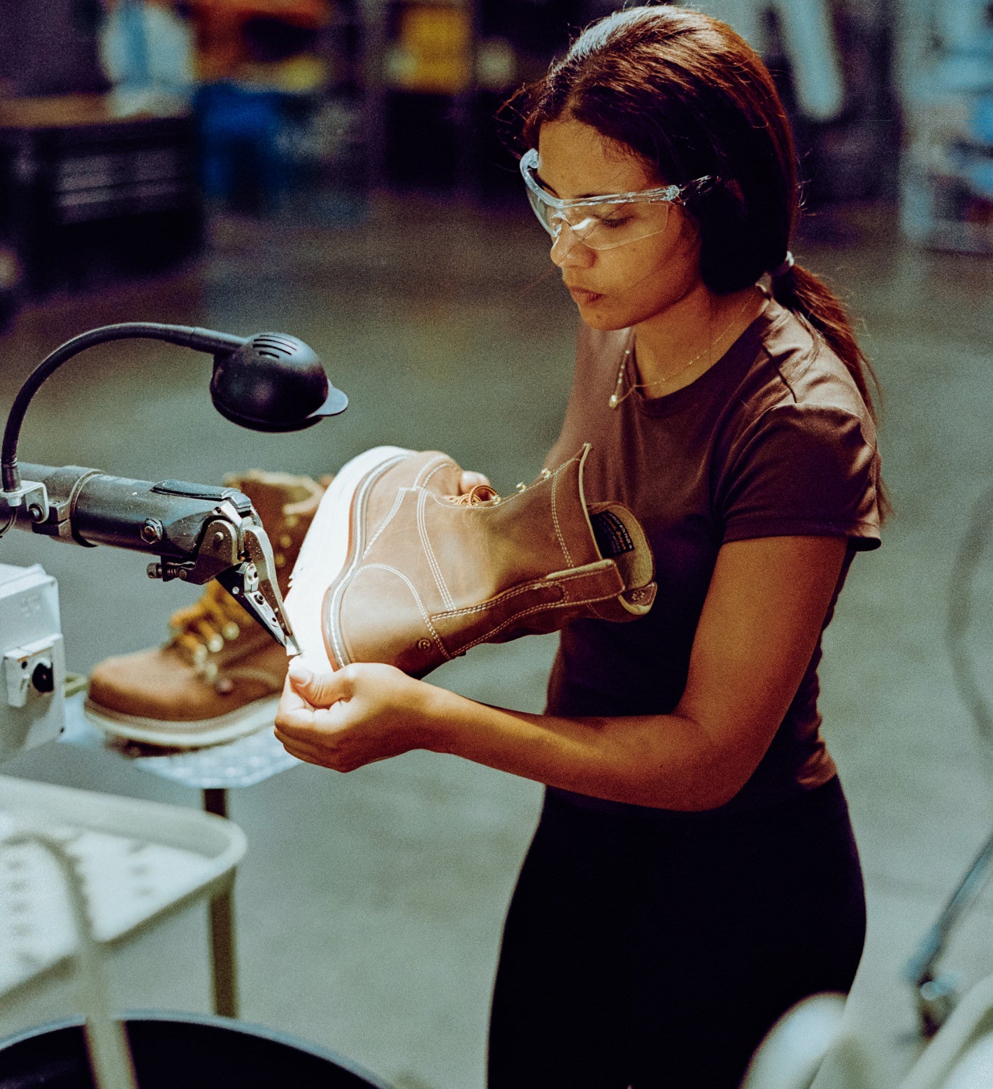 Woman wearing safety glasses holding a brown leather work boot under a machine at a factory workstation.