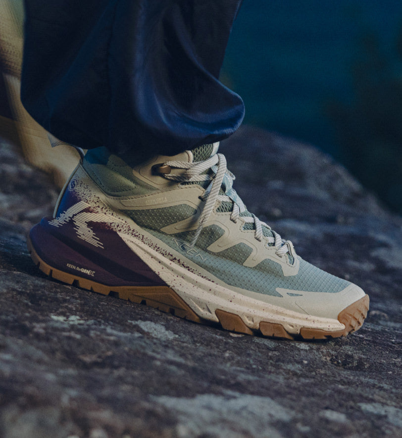  A woman stands on a rock, wearing Women's Targhee Apex boots and sneakers, showcasing new outdoor footwear.