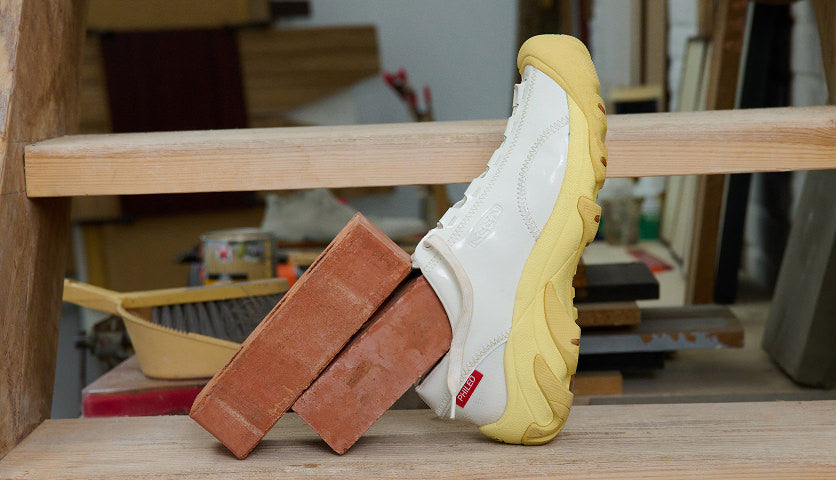 Close up of wooden stairs and two red bricks, one brick is wearing a white KEEN & Phileo collaboration Targher shoe