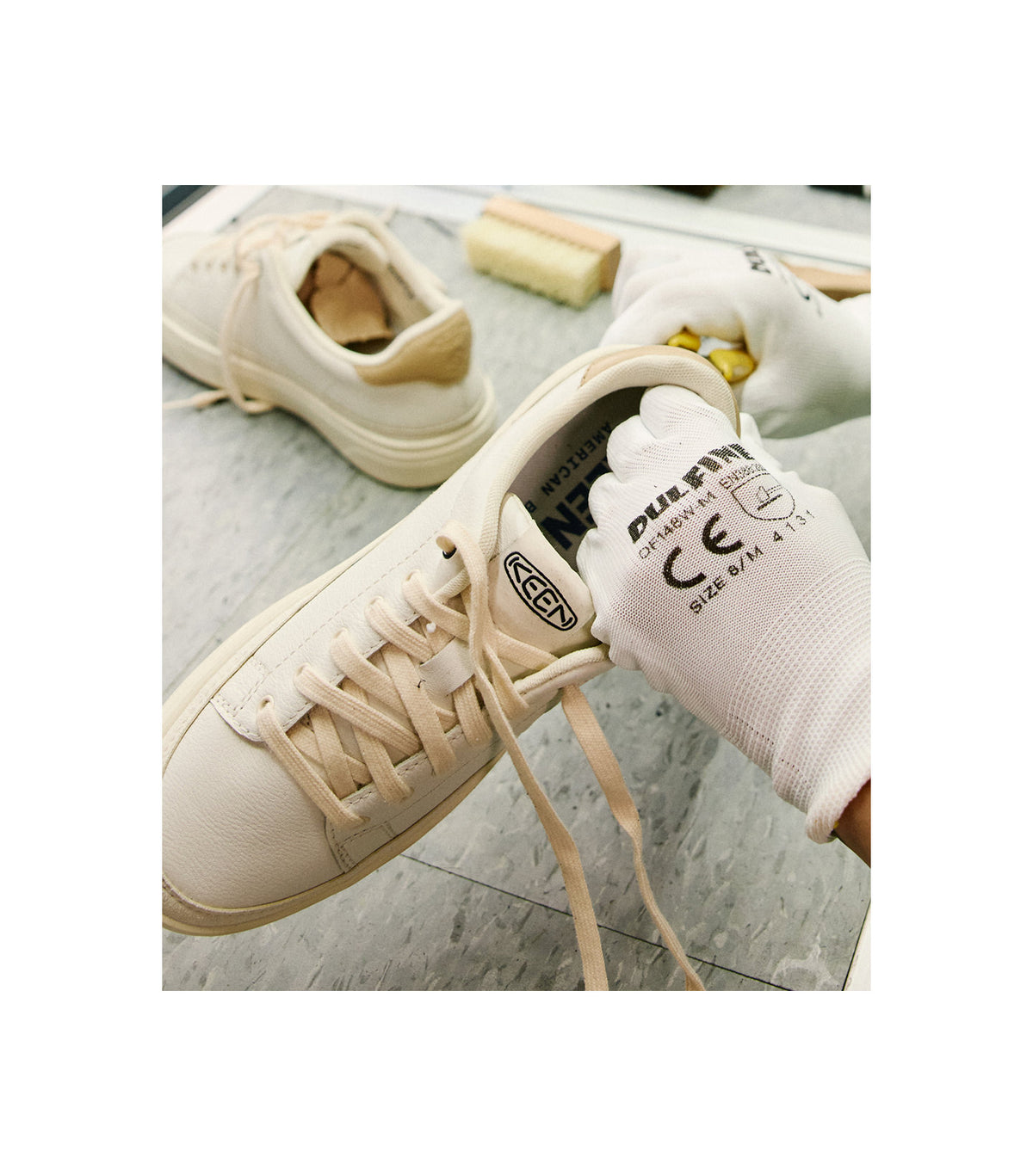 A close up shot of someone in white work gloves using yellow pliers to attach a leather piece to white and tan Keen Made sneakers on a factory counter. 