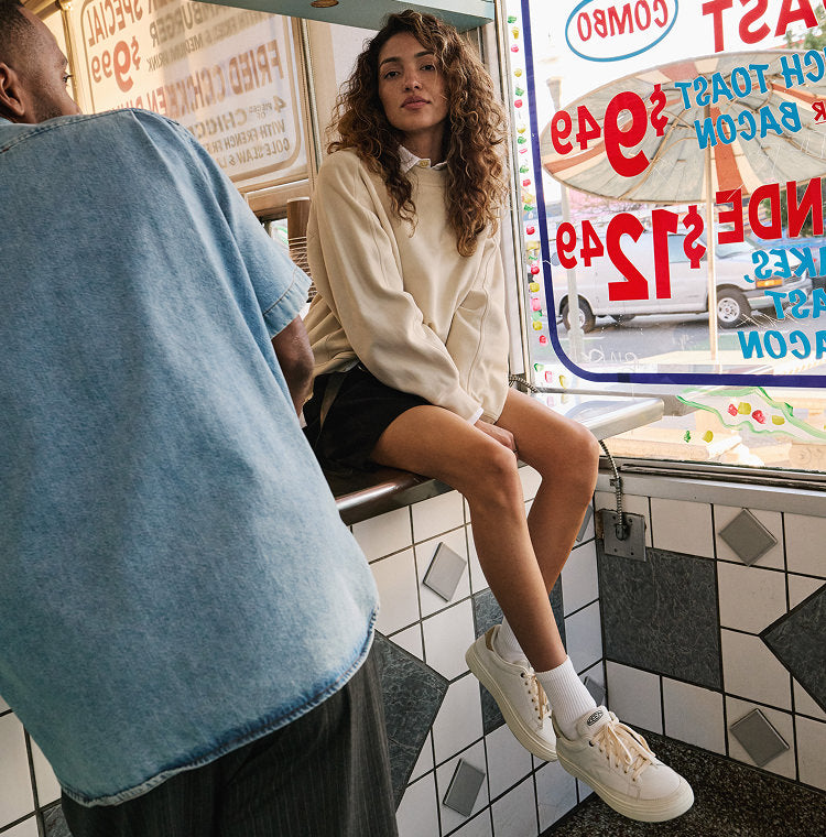 A fashionable woman sitting on the counter of an old school taco shop while wearing a white collared sweater and white Keen Made sneakers. 