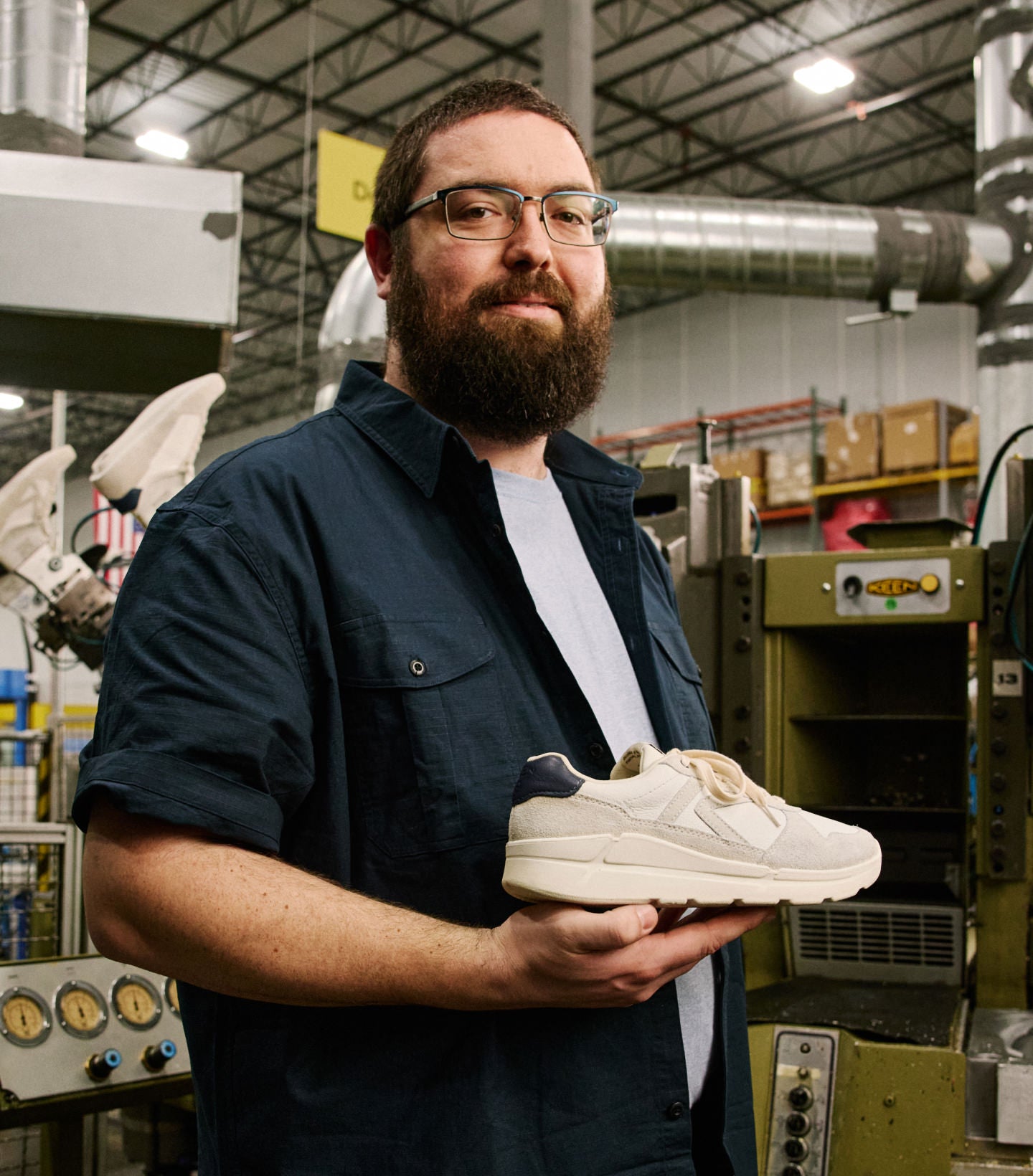 A bearded man with glasses holding a KEEN made leather sneaker at the KEEN factory with production materials  and machines in the background. 