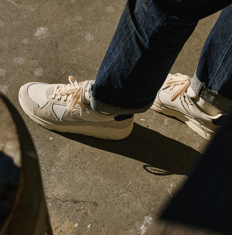 A man in blue jeans an white and tan leather sneakers walking across city street. 