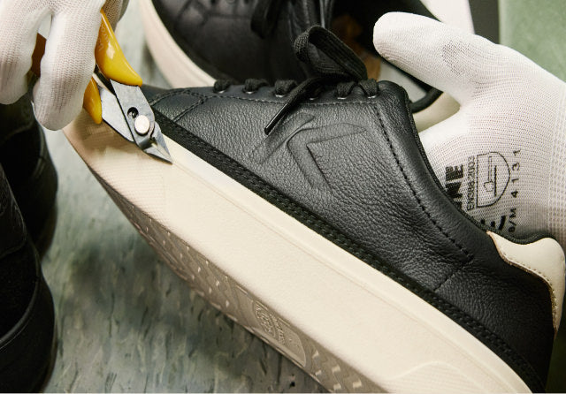 Close up of a person in white gloves using a small metal tool to perfect the white sole of a black and white leather sneaker in a factory setting. 
