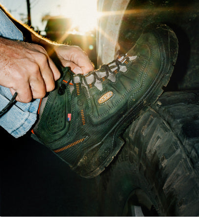 Man with his foot on a tire, tying his dark green Targhee IV work boot.