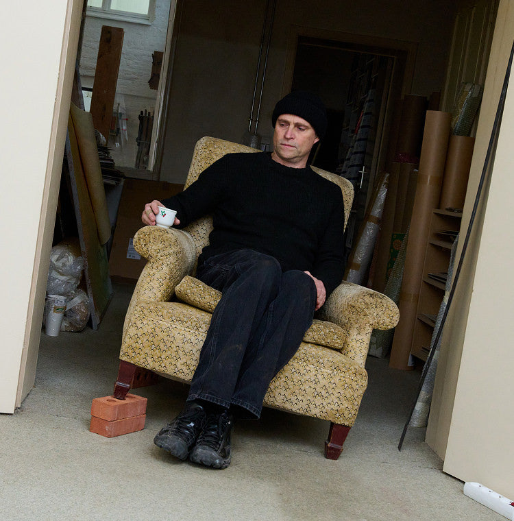 Artist sitting in dusty cushioned chair with espresso cup and wearing black Targher shoes