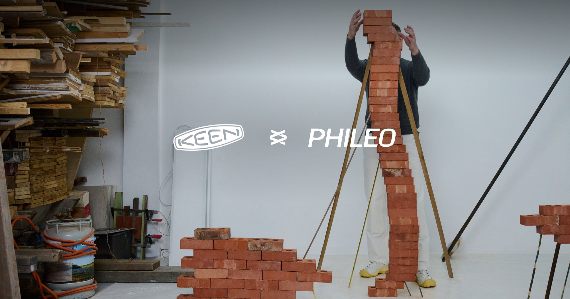 Philéo Landowski, stands in his studio behind red bricks he is stacking in a snaking pattern taller than himself while wearing the Targher collaboration shoes