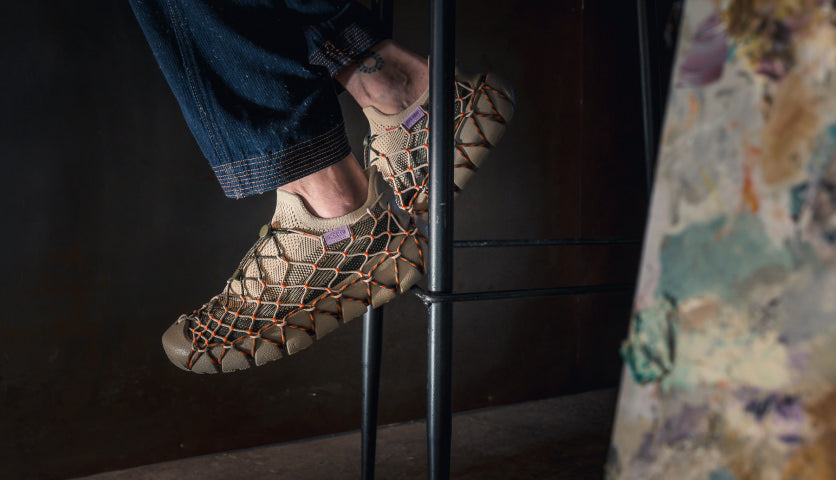 A man's feet on a bar stool, wearing webbed Uneek 360 sneakers in a special collaboration with Henrik Vibskov.