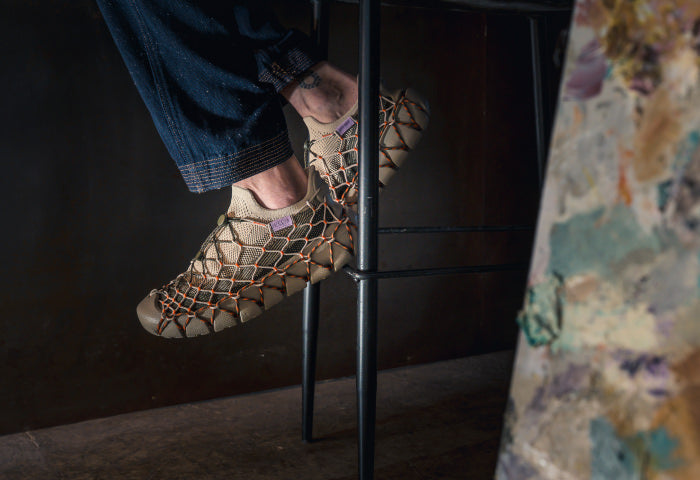 A man's feet on a bar stool, wearing webbed Uneek 360 sneakers in a special collaboration with Henrik Vibskov.