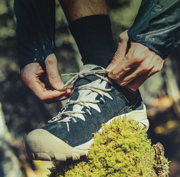 A man tying the laces to his hiking shoes while his foot is perched on top a mossy rock. 