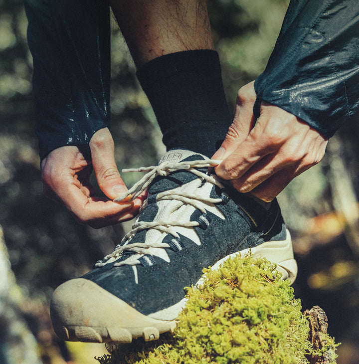 A man tying the laces to his hiking shoes while his foot is perched on top a mossy rock. 