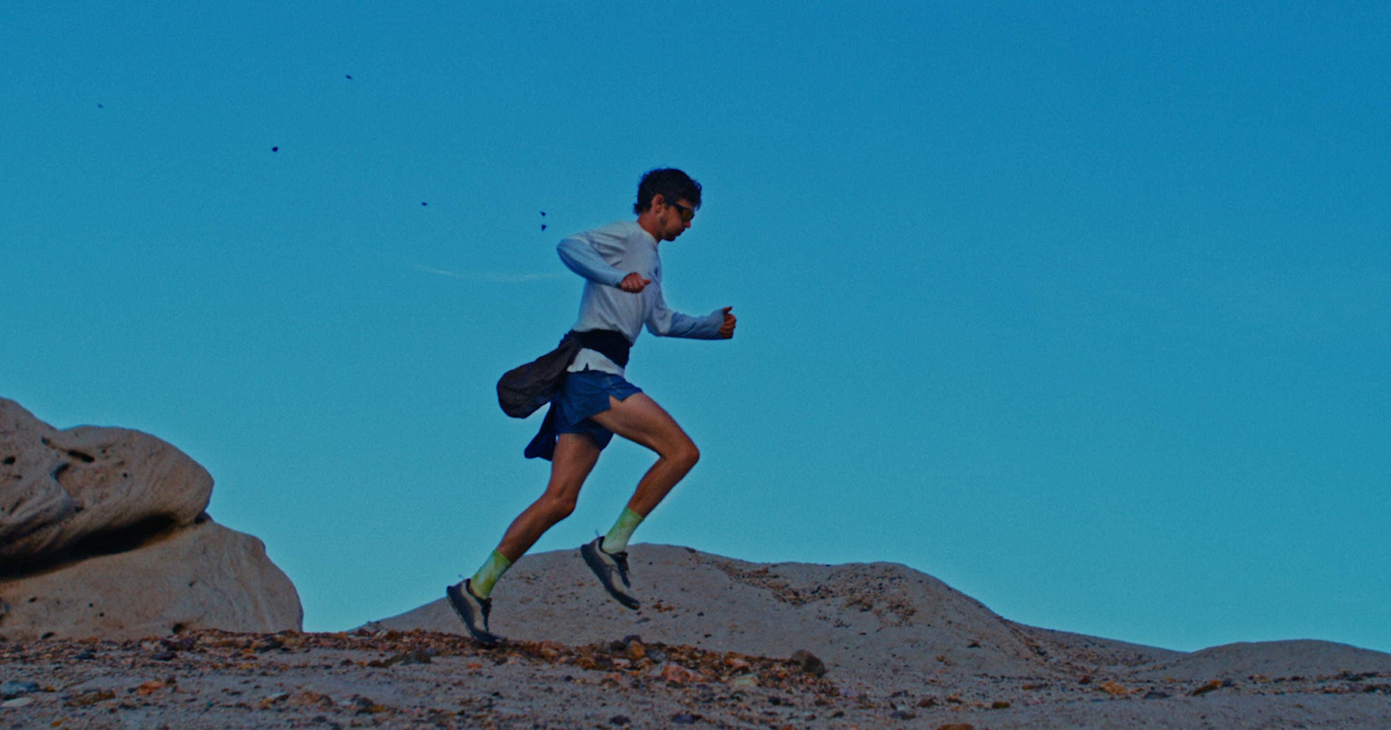 A trail runner in very early morning light, running over desert terrain while wearing Wander running shoes. 