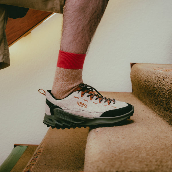 A man in red and orange crew socks wearing Jasper Zionic shoes in tan, orange, black and dark green while walking up carpeted stairs. 