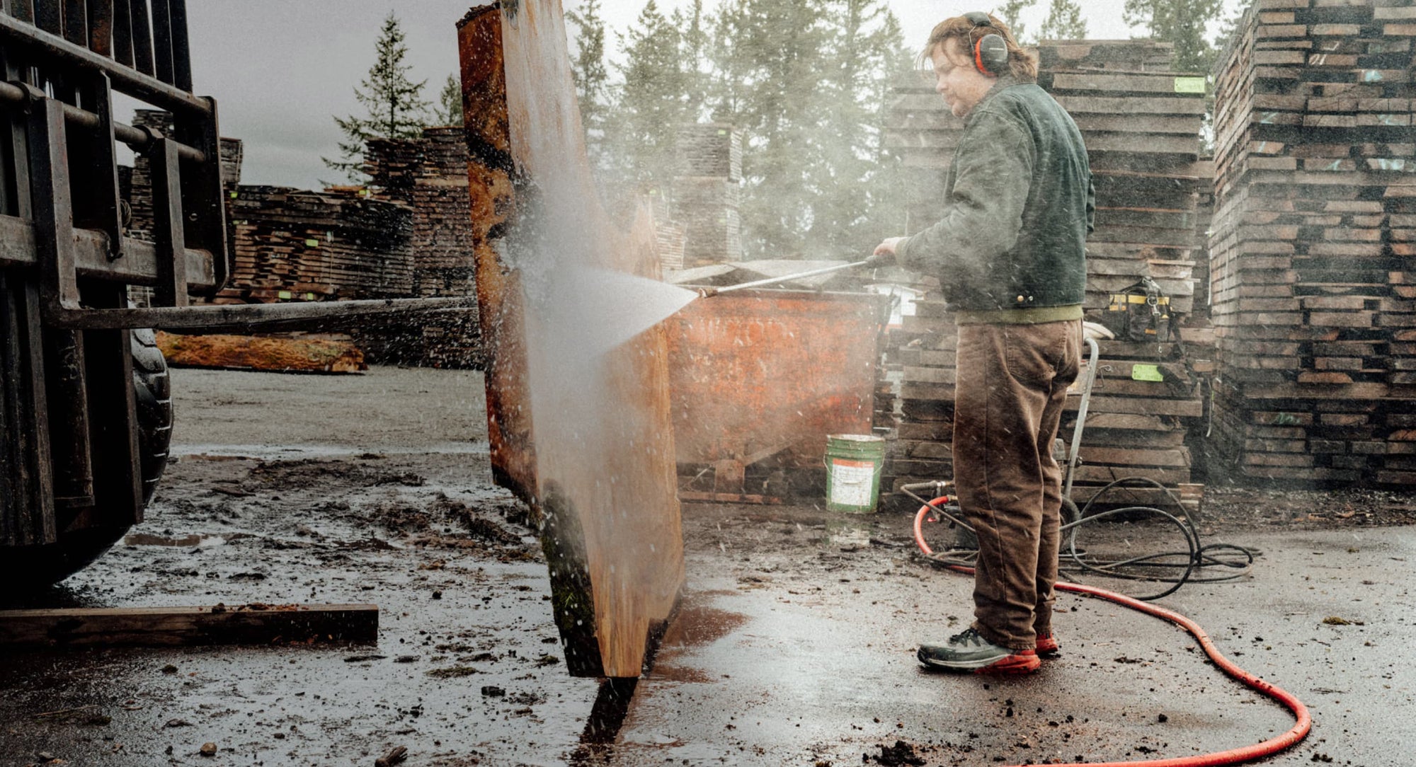 A man with ear protection power washing a giant slab of wood in a lumberyard while wearing waterproof Targhee Blur work boots.