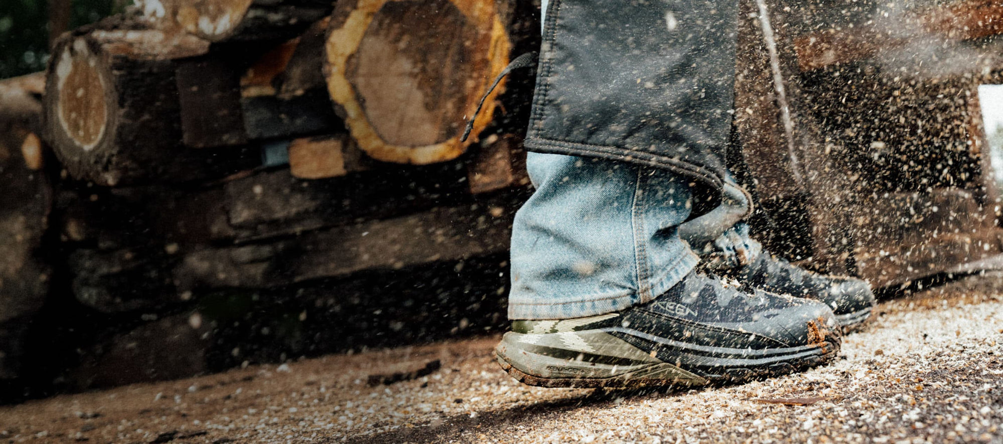 A man in jeans and protective coverings wearing Targhee Blur utility boots with wood chips flying around his feet. 