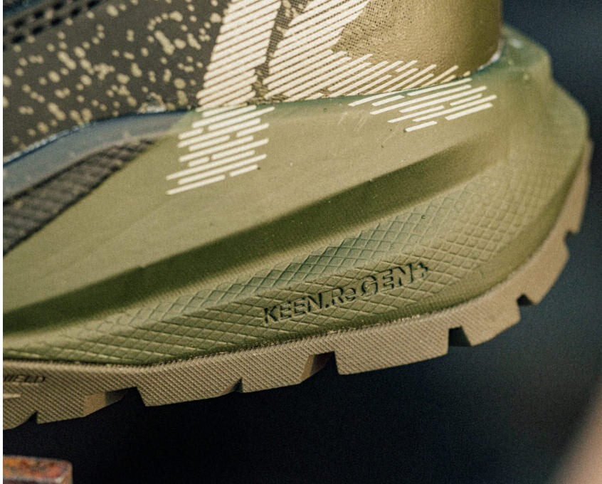 A pair of Targhee Blur work boots in dark green and black being worn on rocky dirt ground, showcasing the lightweight and compression-resistant midsole. 