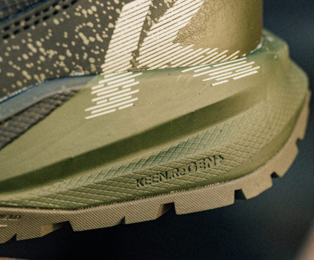 A pair of Targhee Blur work boots in dark green and black being worn on rocky dirt ground, showcasing the lightweight and compression-resistant midsole. 