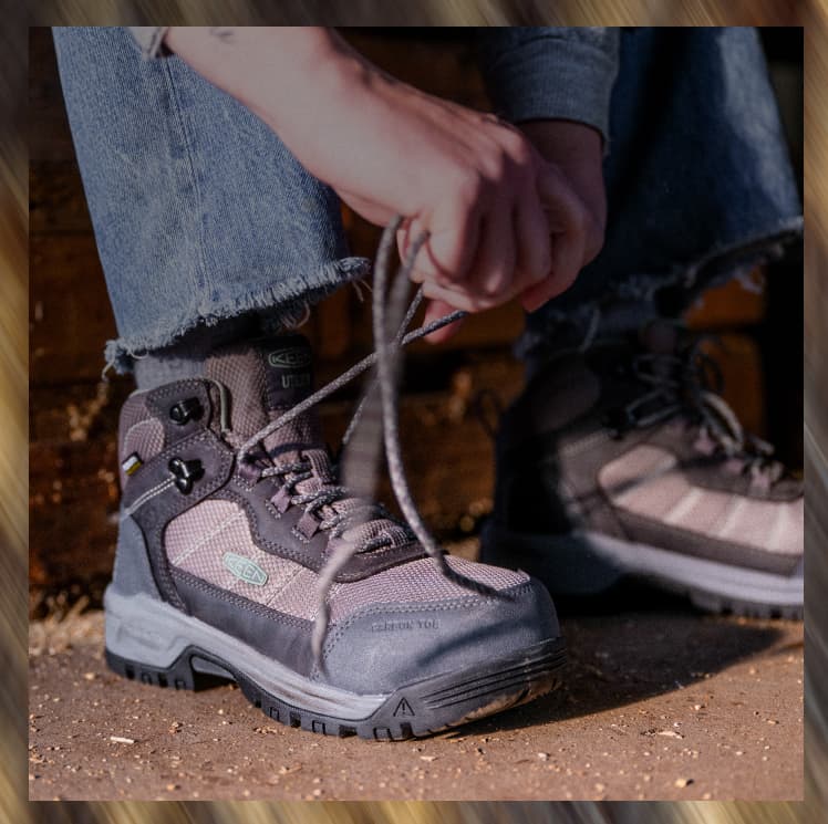 A woman sitting on a wood block lacing up her grey, waterproof Skokie work boots with a hiking-boot aesthetic and carbon toe. 