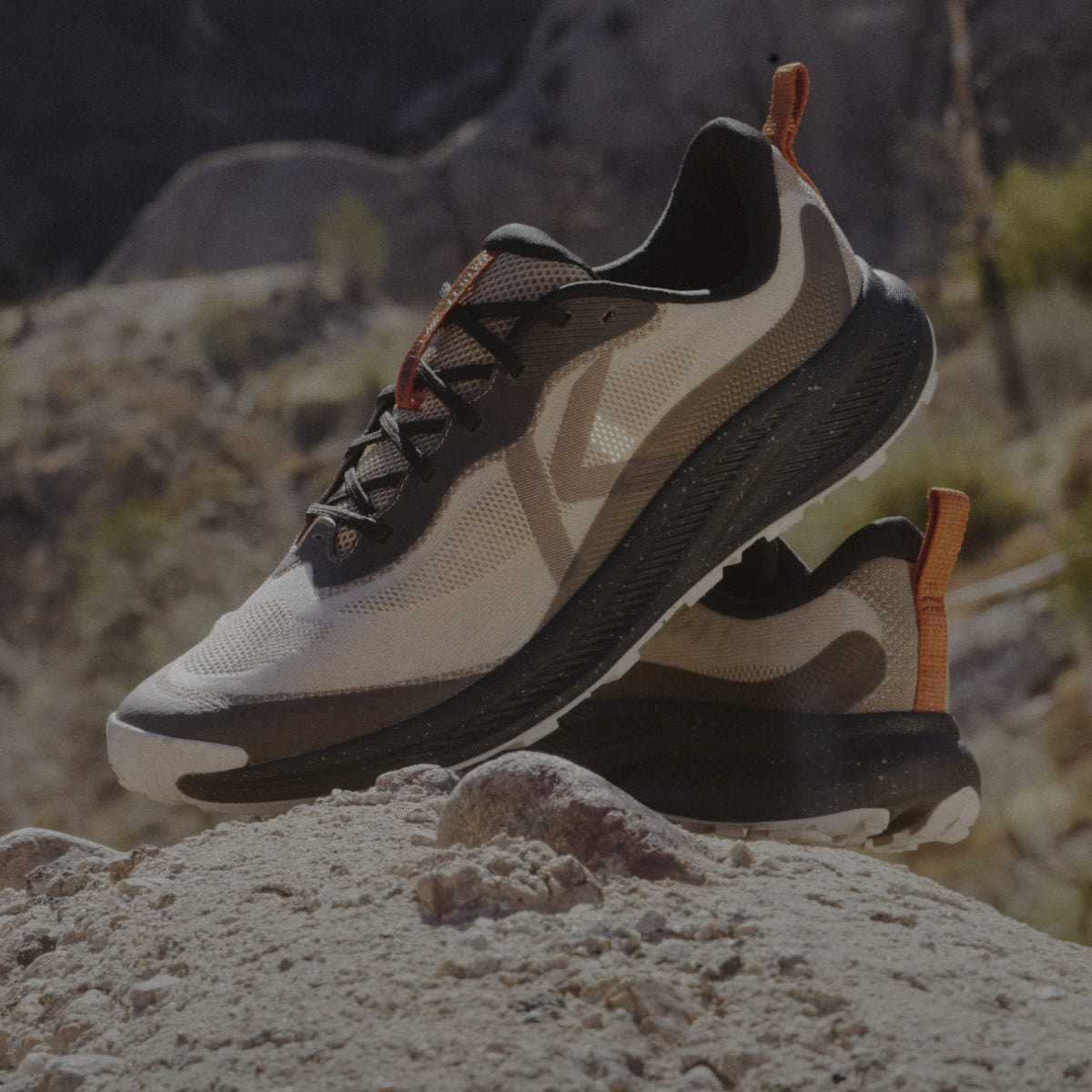 A pair of men's Wander running shoes in  tan and black with orange detail posed on a desert rock. 