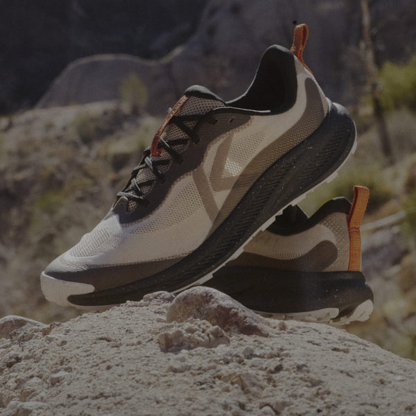 A pair of men's Wander running shoes in  tan and black with orange detail posed on a desert rock. 