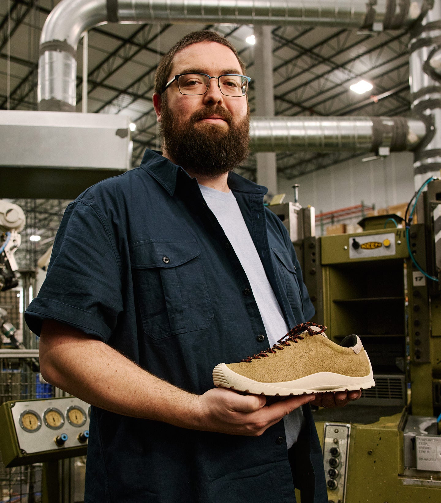 A bearded man with glasses holding a KEEN made leather sneaker at the KEEN factory with production materials  and machines in the background. 