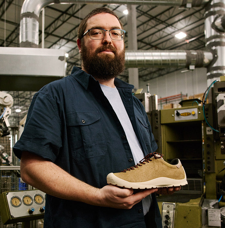 A bearded man with glasses holding a KEEN made leather sneaker at the KEEN factory with production materials  and machines in the background. 