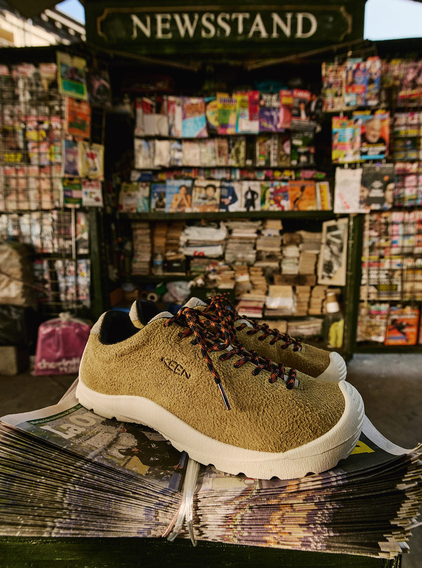 A pair of men's Jasper Lite sneakers in tan leather suede with red and black laces placed on a stack of newspapers with a newsstand in the background. 