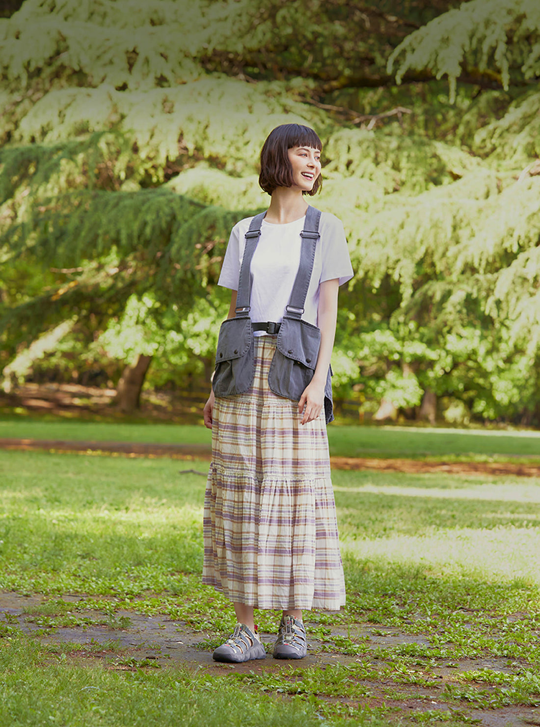 A woman in a long plaid skirt and stylish vest wearing the Fuji Rock collaboration hyperport sandals while smiling in the park.