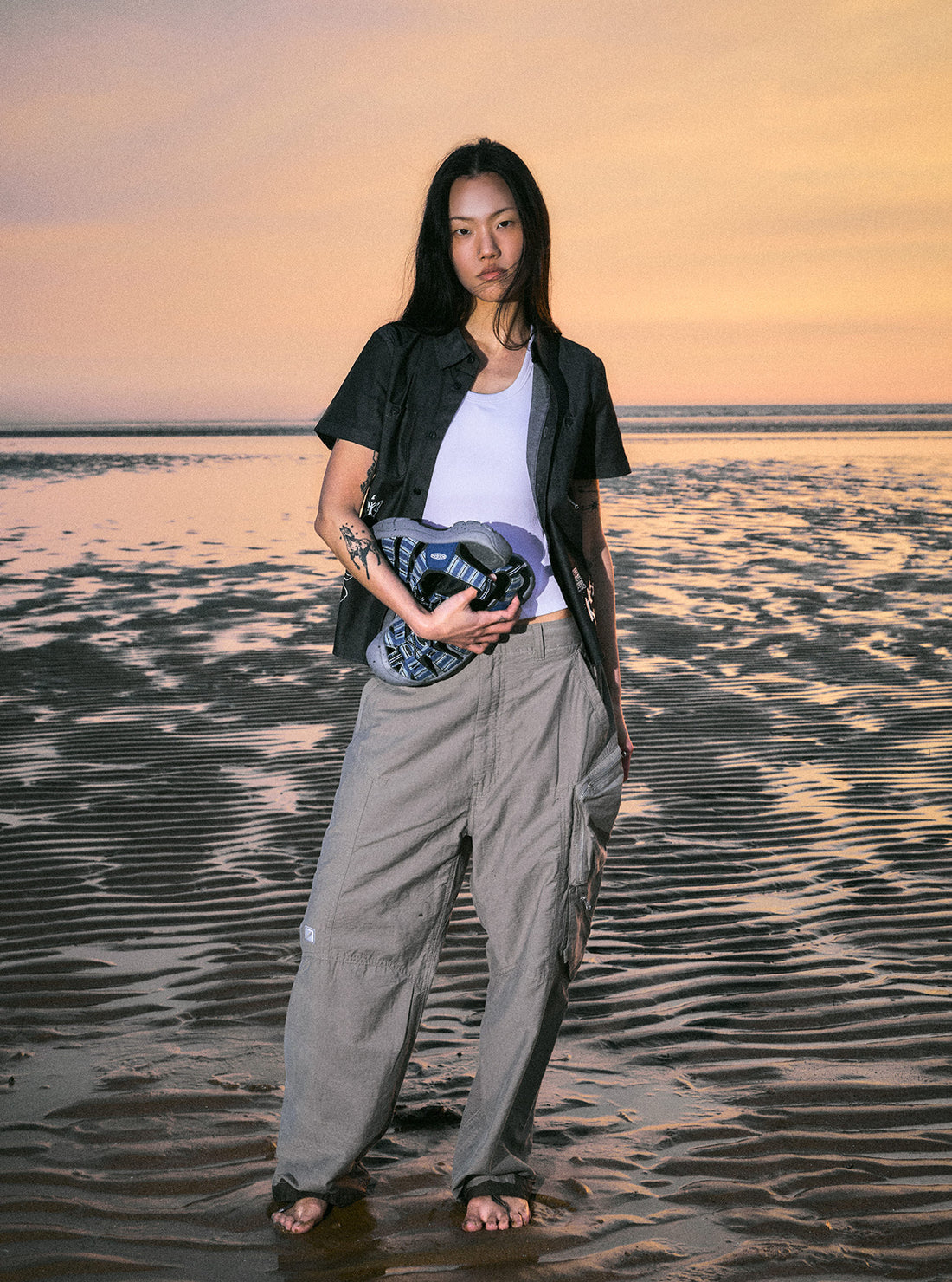 A woman standing  barefoot on the beach at sunset while holding YESEYESEE  and KEEN collaboration sandals in her hand.