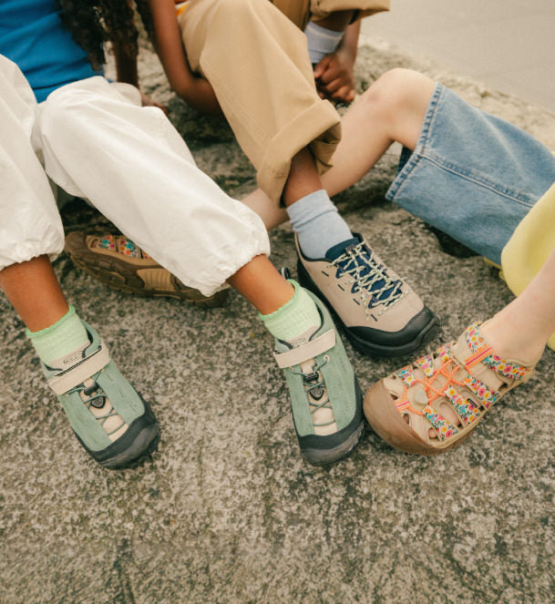 3 pairs of kids feet with keen sneakers and closed toed sandals sitting on a rock.