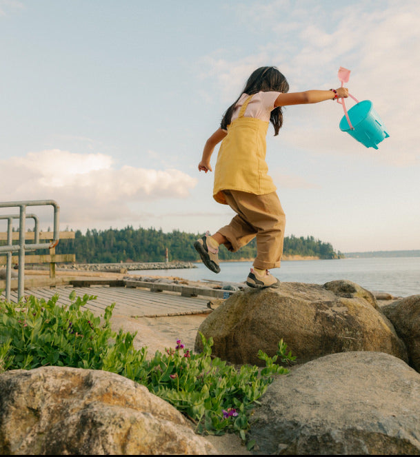 Little girl jumping from boulder to boulder at the beach carrying a bucket wearing KEEN kids' Jasper shoes