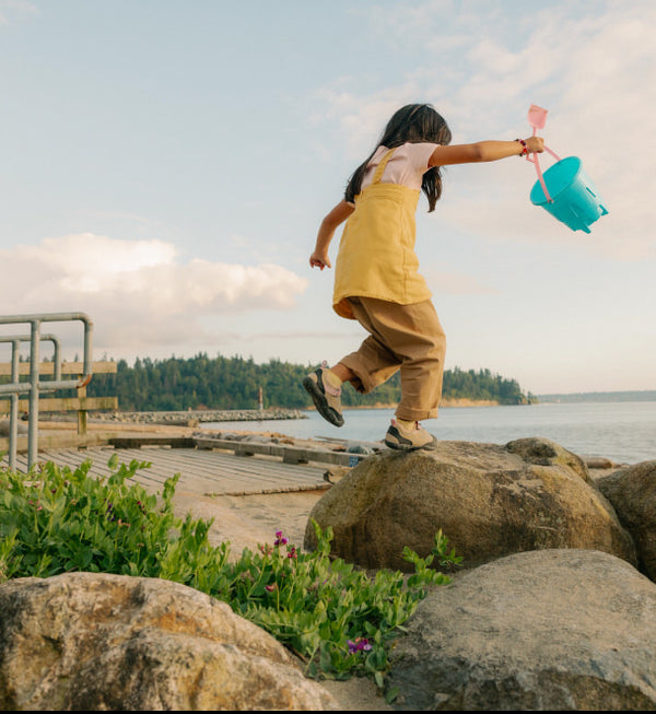 Little girl jumping from boulder to boulder at the beach carrying a bucket wearing KEEN kids' Jasper shoes