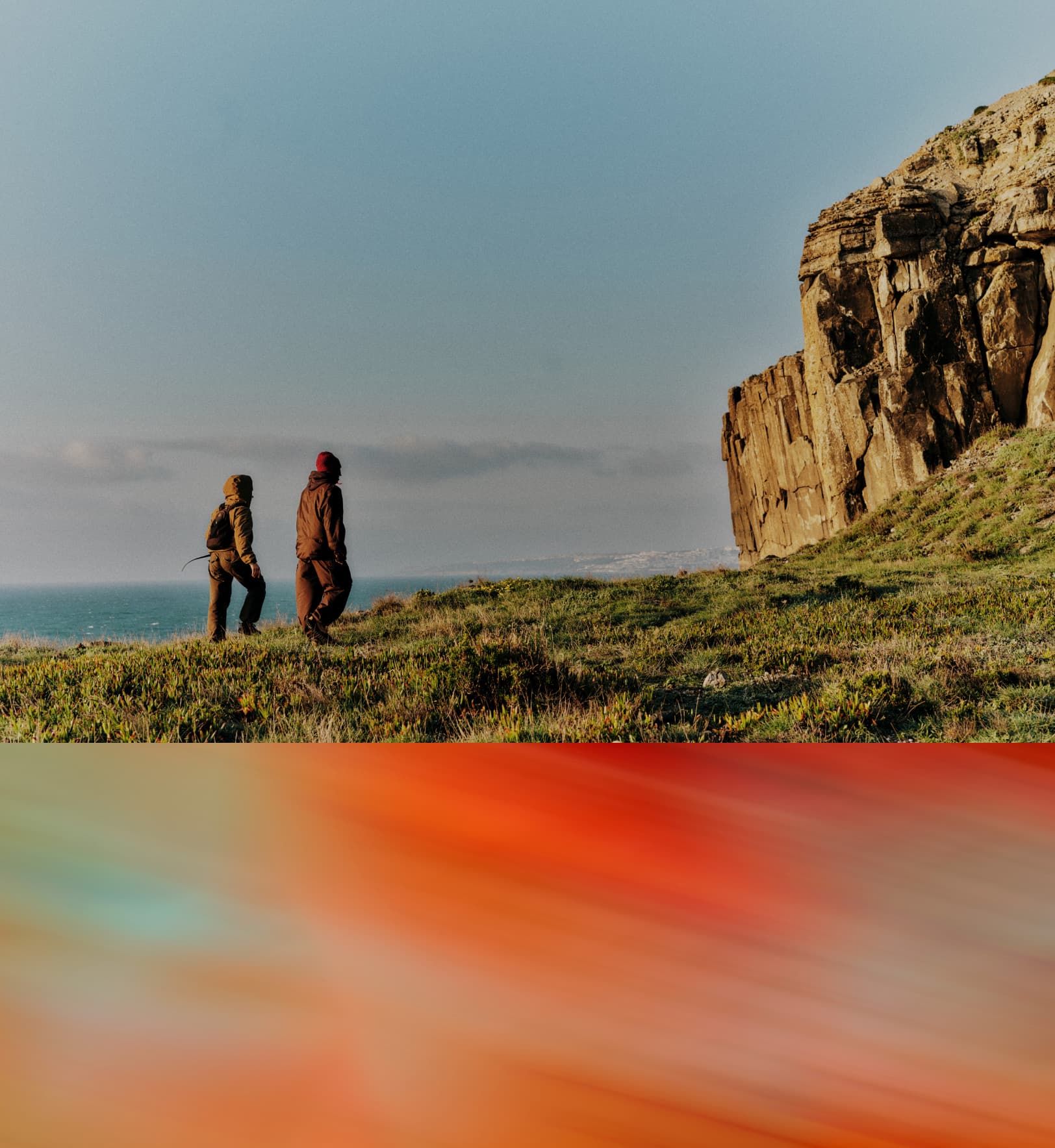 Two people standing on a grassy ledge overlooking sandstone cliffs near ocean waters.