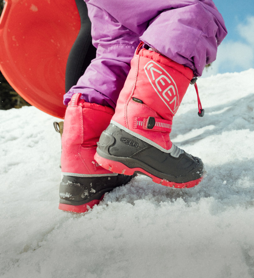 Knee down shot of young girl carrying sled and wearing Snow Troll winter boots while walking up snowy hill. 