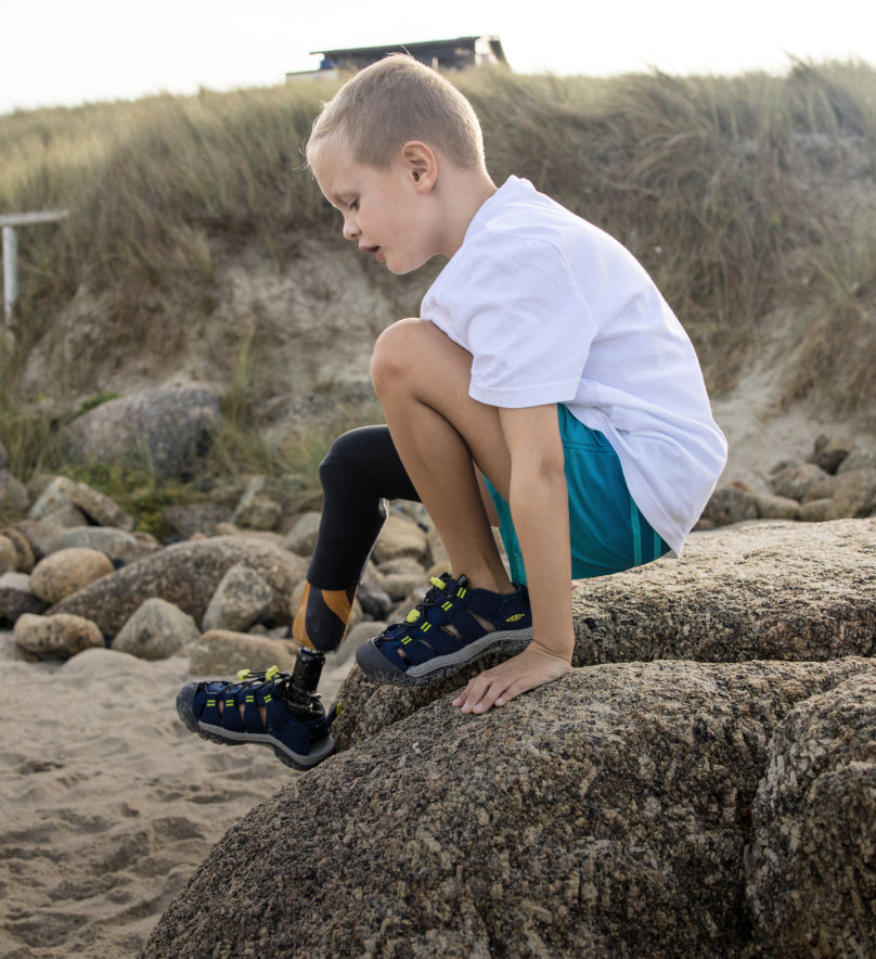 Young boy with a bionic leg wearing Newport Boundless sandals and crawling over large beach boulder. 